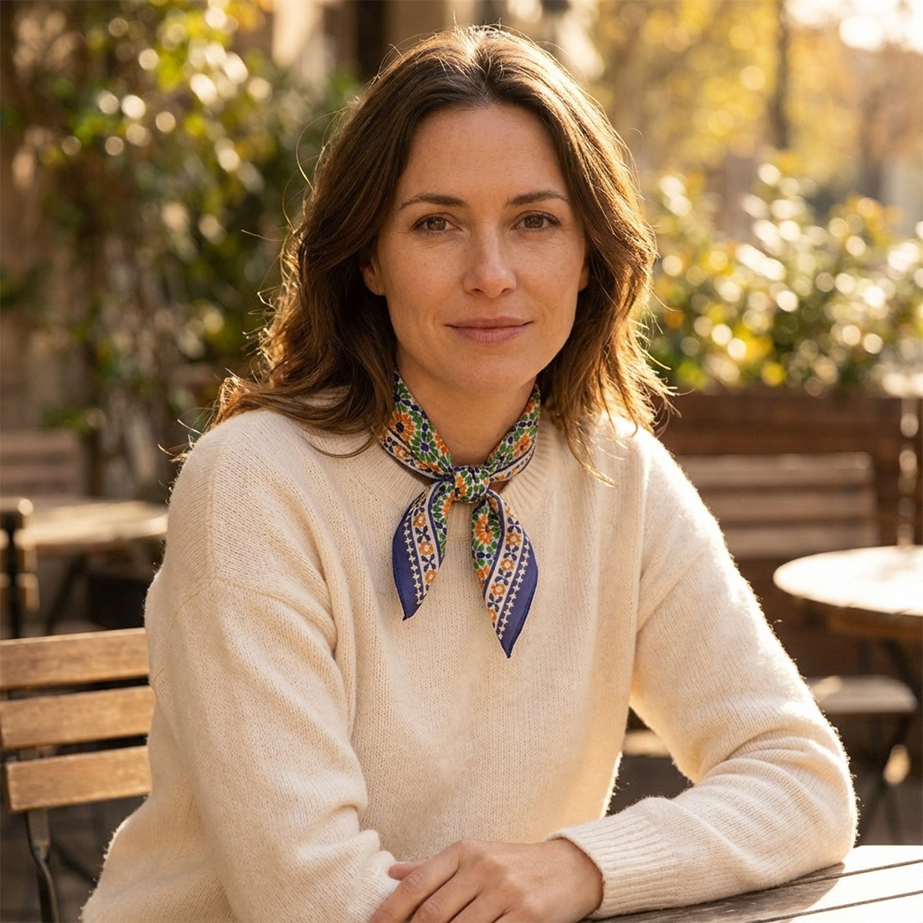A woman with wavy brown hair sits at an outdoor café table, wearing a cream sweater and the Islamic Art Print Square Silk Scarf Nur around her neck. She smiles softly, surrounded by greenery and sunlight.