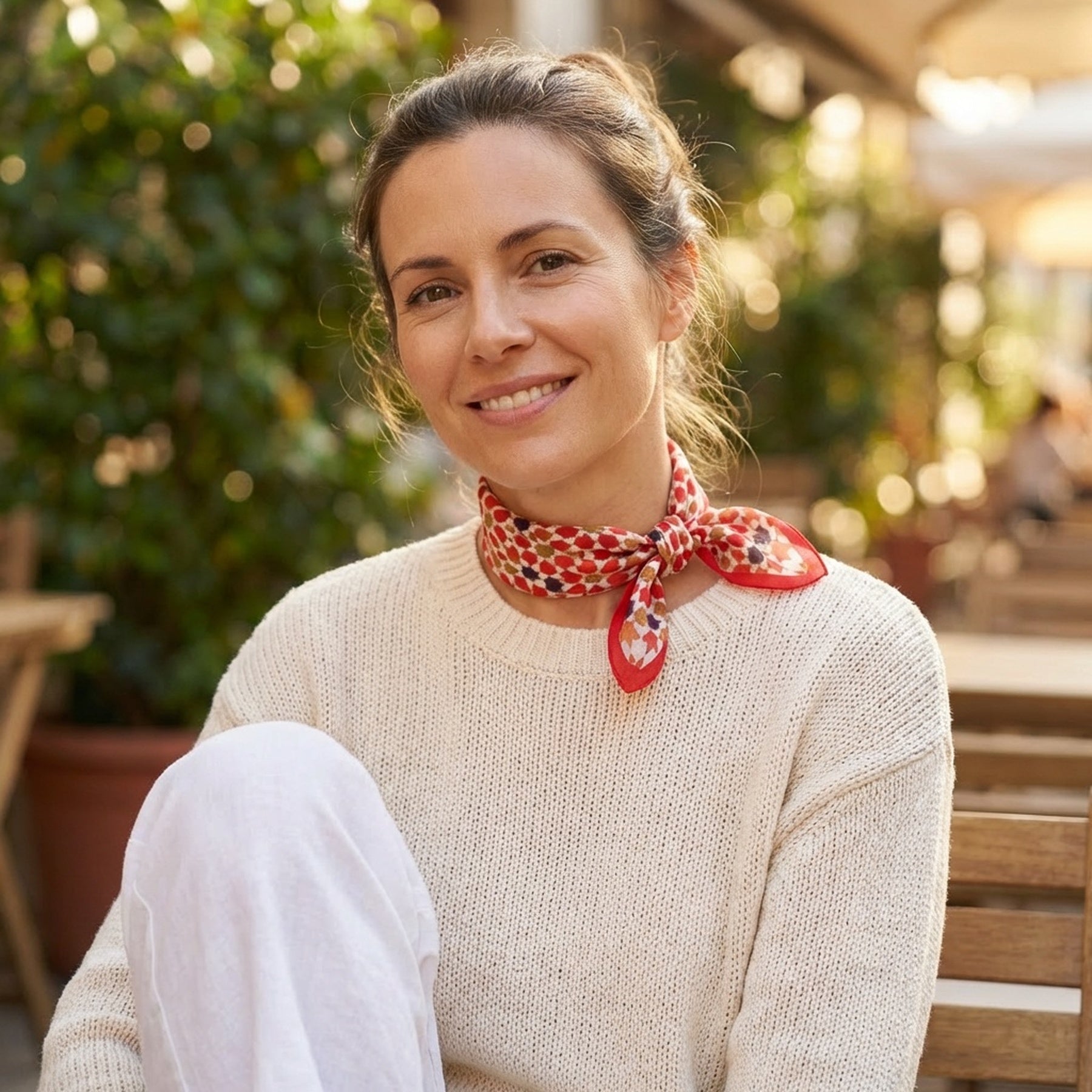 A woman with brown hair tied back, in a cream sweater, white pants, and the Small Square Scarf Alhambra—featuring an Islamic art print—smiles at the camera while sitting outdoors on a wooden chair among green plants and tables.
