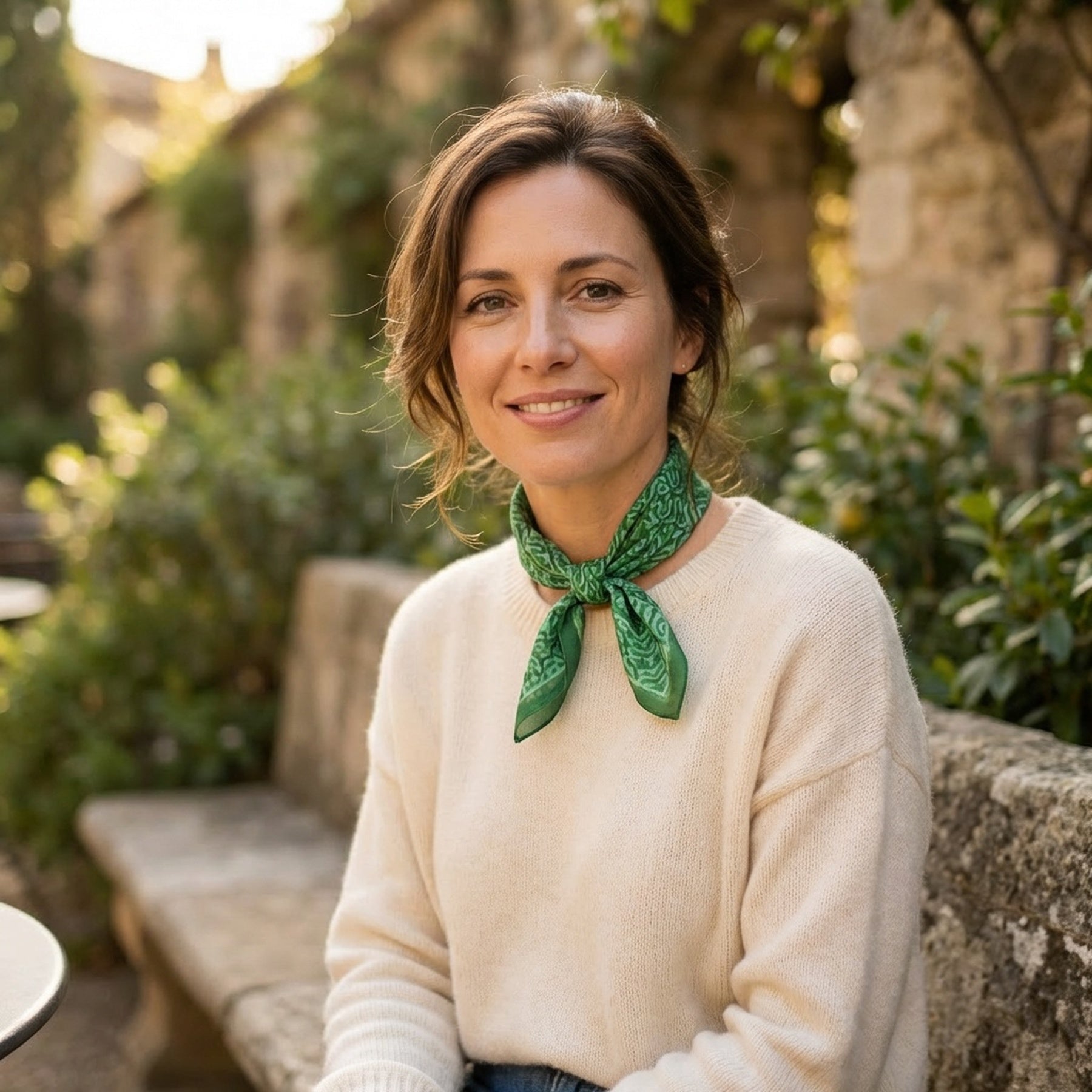 A woman with brown hair in a loose bun, wearing a cream sweater and the Green Square Silk Scarf Fihri with an Islamic art print, sits smiling on a stone bench at the Alhambra Palace, surrounded by sunlit gardens and historic stone walls.