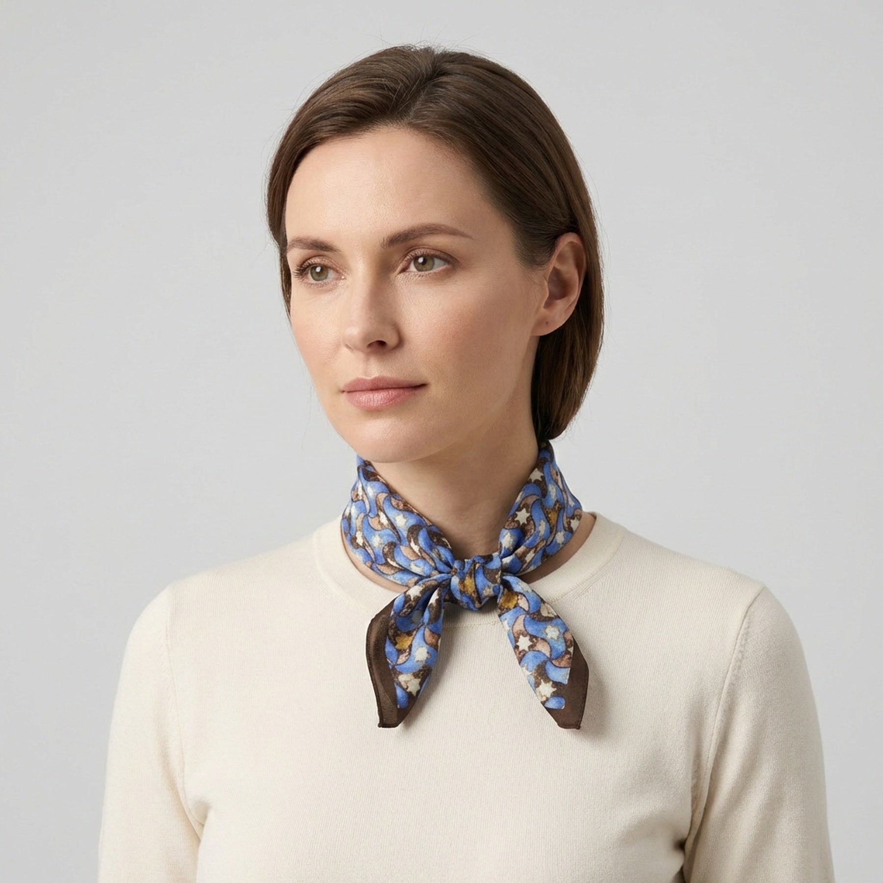 A woman with light brown hair wears the Silk Square Scarf for Women Brown, tied at her neck over a cream sweater, evoking Spanish craftsmanship, as she looks slightly to the side against a plain light gray background.