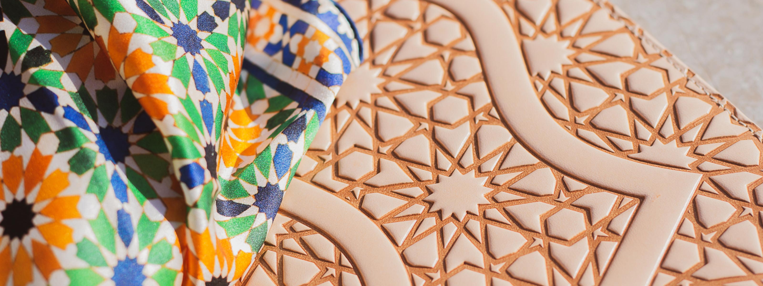A colorful patterned fabric lies on a surface with an intricate geometric design engraved in light brown material, both featuring traditional Islamic art motifs.