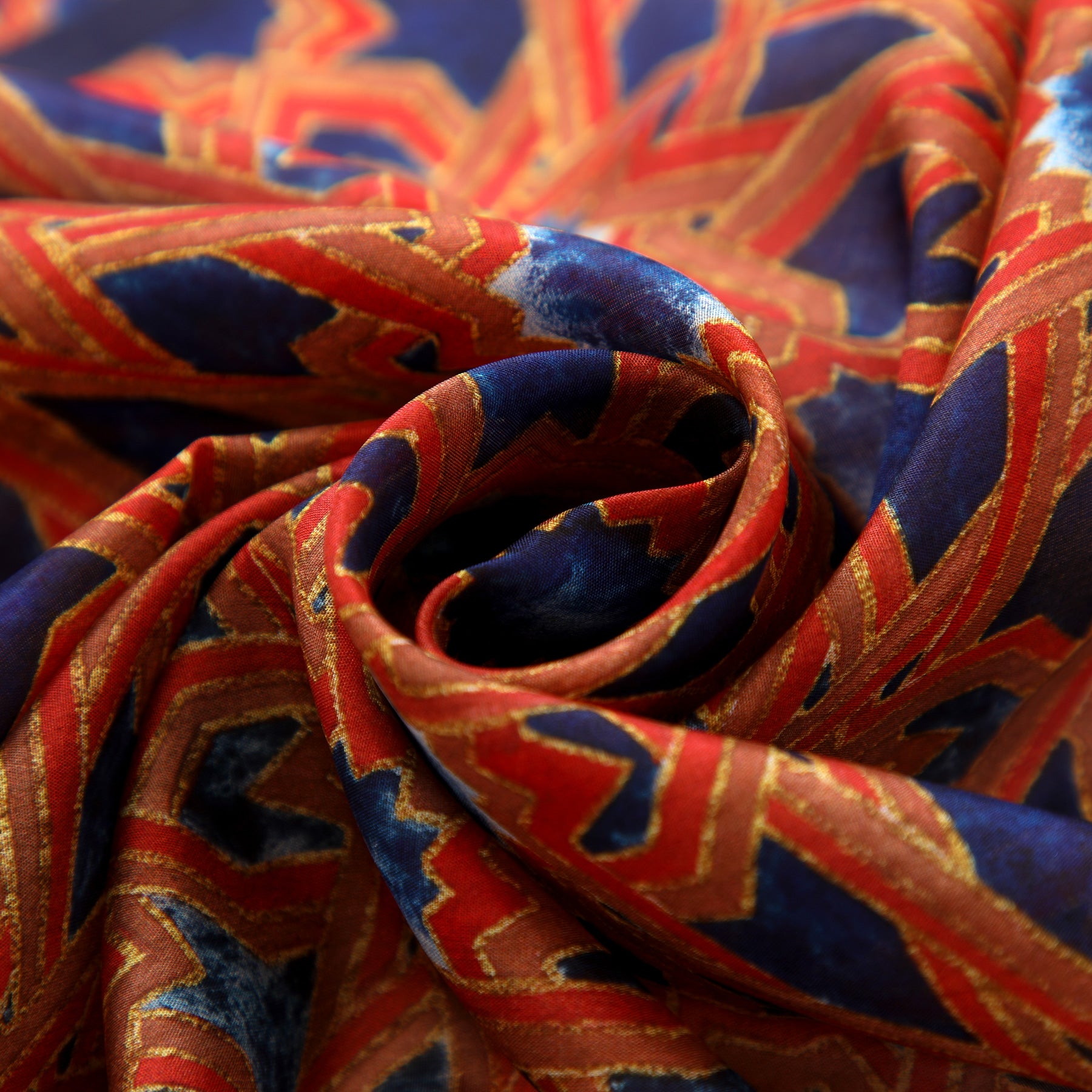 A close-up of the Islamic Art Silk Scarf Alcázar Genil, showcasing vivid red, blue, and gold geometric patterns inspired by Andalusian art, gathered into a soft spiral fold.