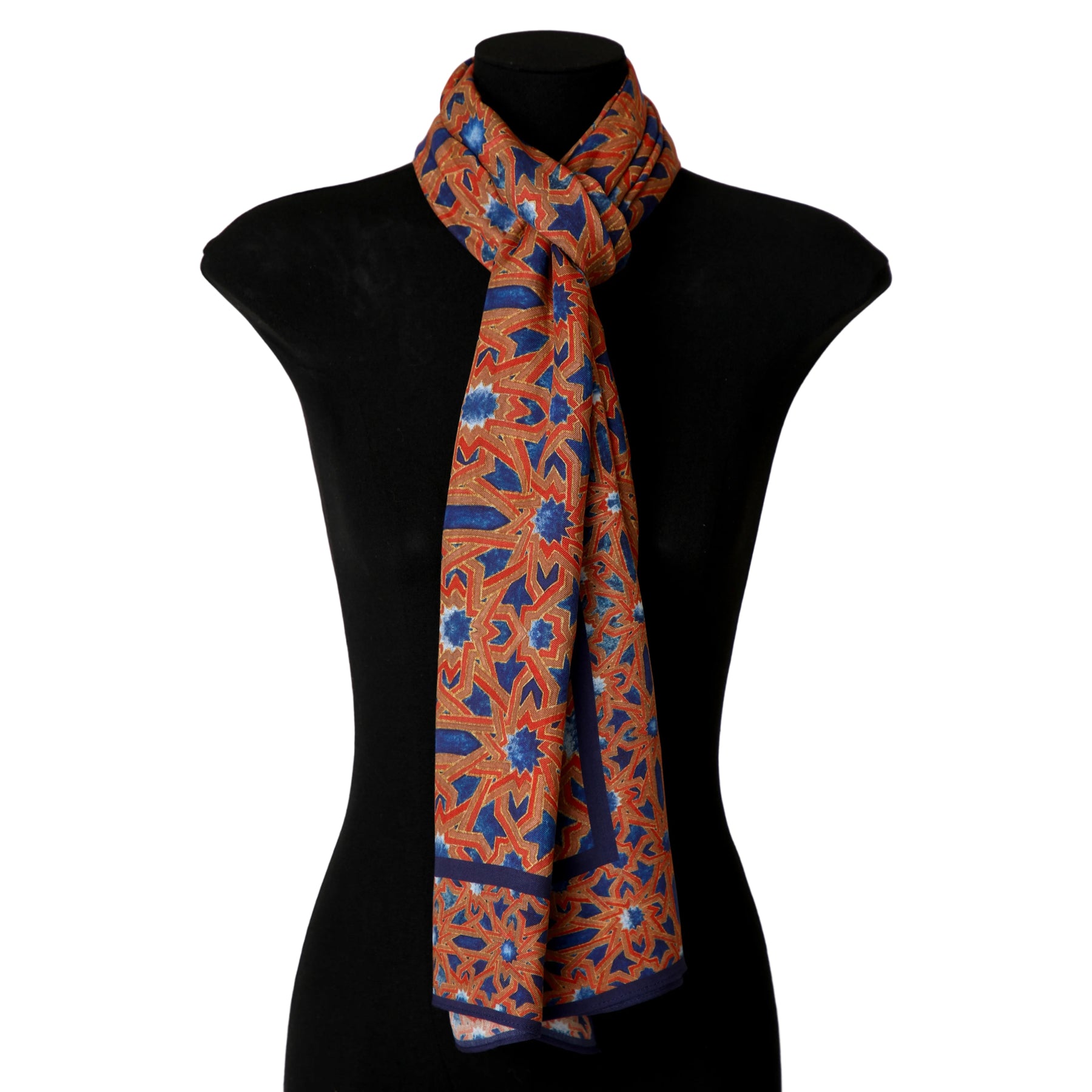 A mannequin showcases the Moorish Tiles Inspired Modal Scarf - Alcázar Genil, featuring an intricate orange, blue, and red geometric pattern reminiscent of Andalusí Islamic art, draped and knotted at the neck against a plain black background.