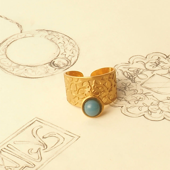 The Platónica Nasrid Palace Inspired Ring Gold & Blue, featuring intricate floral designs and a blue centerpiece, is elegantly displayed on a sheet of pencil jewelry sketches.