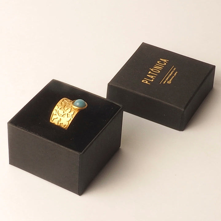 The Nasrid Palace Inspired Ring Gold & Blue by Platónica features an embossed gold design with a turquoise stone, displayed on a black box. Another black Platónica box in gold lettering sits nearby on a light background.