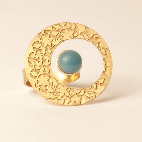 The Platónica Alhambra Palace-Inspired Gold Ring for Women features an intricate floral design with a circular shape and open center, highlighted by a single polished blue glass stone set near the edge against a plain light cream background.