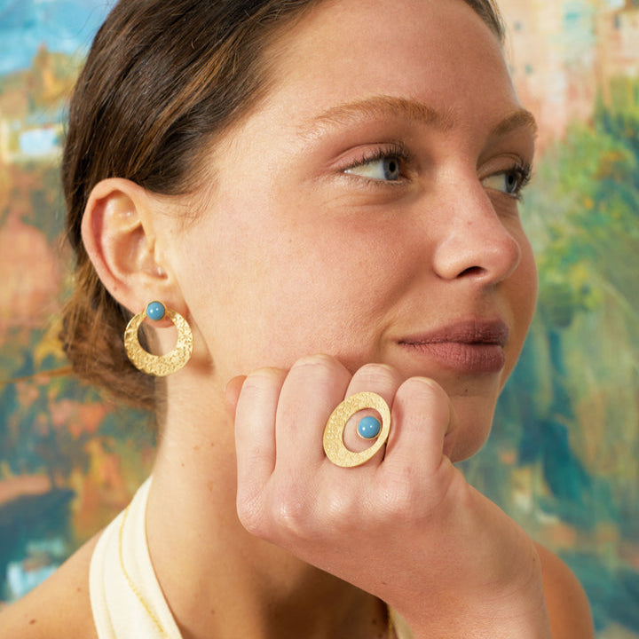 A woman in a cream halter top rests her chin on a hand adorned with Platónicas Alhambra Palace-Inspired Gold Ring with blue detail. Her large circular earrings echo the rings design. A blurred, colorful painting sets the backdrop.