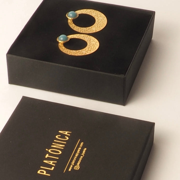 Alhambra Tiles inspired gold earrings with blue stone accents by Platónica, featuring textured hoops, displayed on a black box above another box with PLATÓNICA in gold lettering.