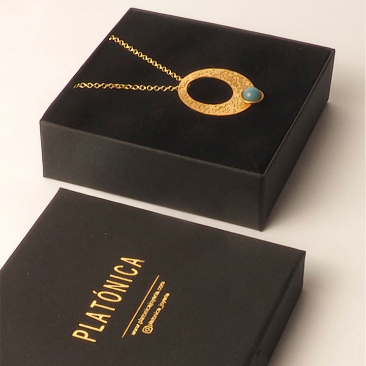 The Alhambra Palace Gold Circle Pendant with Blue Stone by Platónica rests in a black jewelry box. Another black box with PLATÓNICA in gold letters is placed beside it.