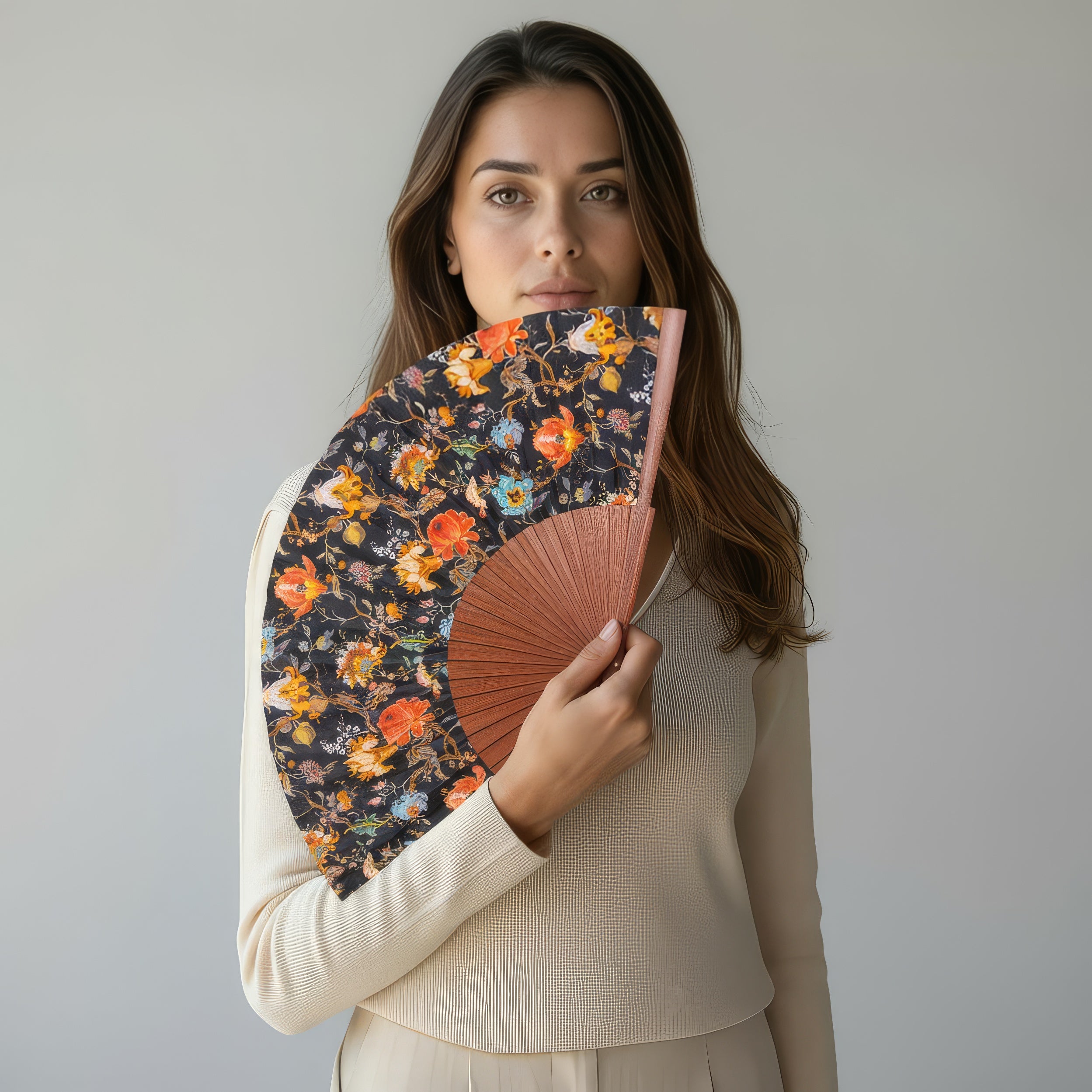 A woman with long brown hair in a cream outfit holds the William Morris Print Hand Fan Aubrey, its vibrant floral design partially covering her face, set against a plain light gray background.
