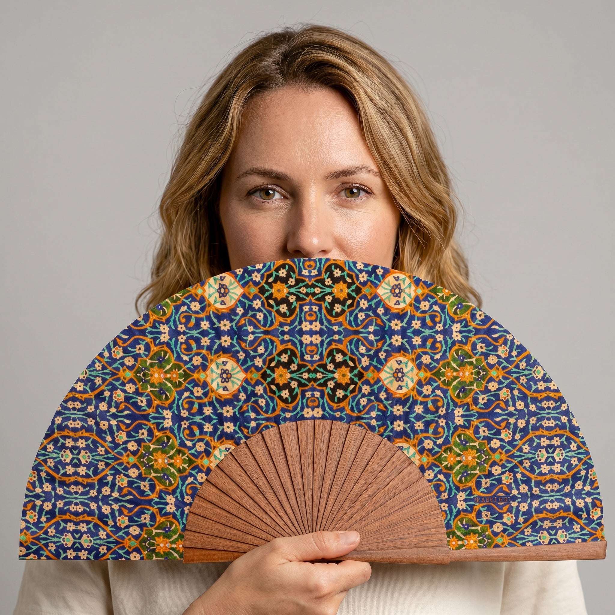A woman holding the Andalusian Mosaic Hand Fan Itr, with its Andalusian mosaic geometric print in blue and orange, against a soft neutral background.