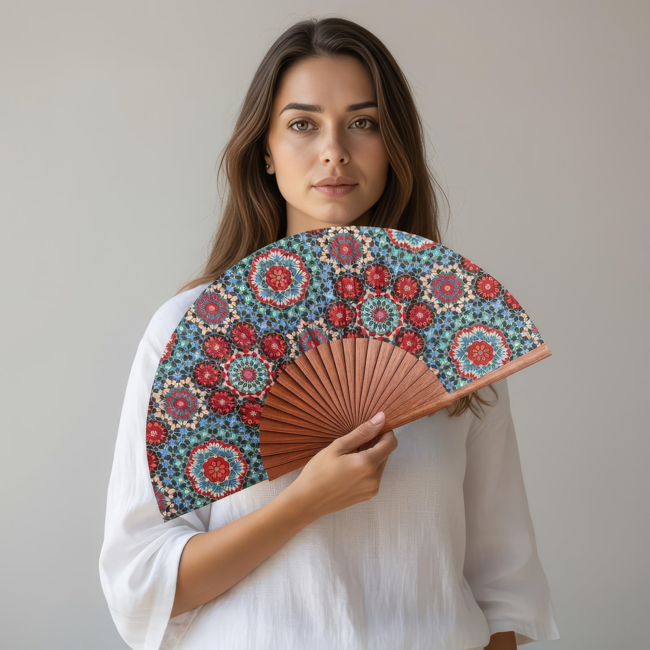 A woman with long brown hair, dressed in a white blouse, holds the Folding Silk Fan Baraka V1—adorned with vibrant Islamic geometric patterns—against her chest, standing before a plain light gray background.