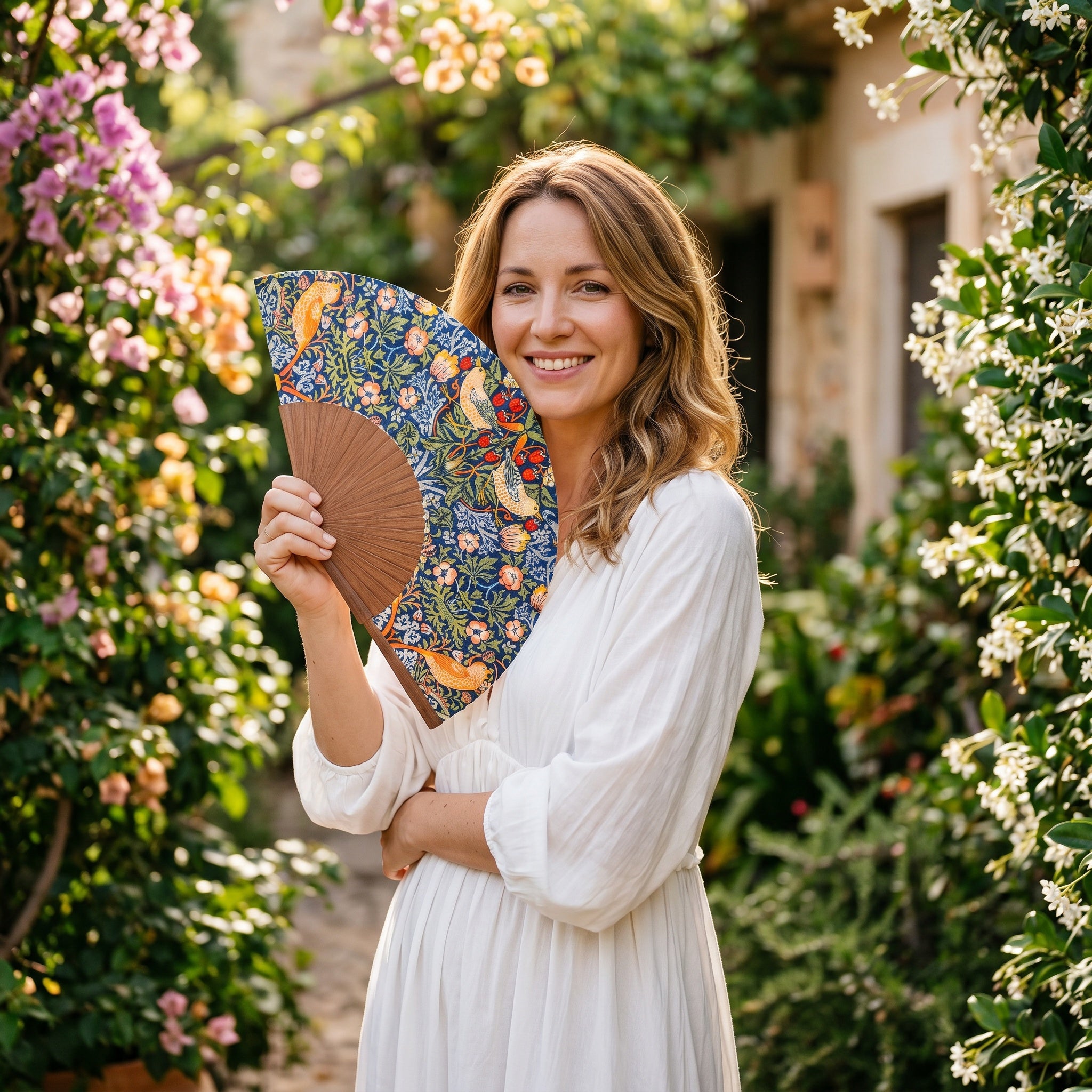 A woman in a summer outfit holding the open William Morris Print Hand Fan Birds II with its William Morris Strawberry Thief print on dark silk.