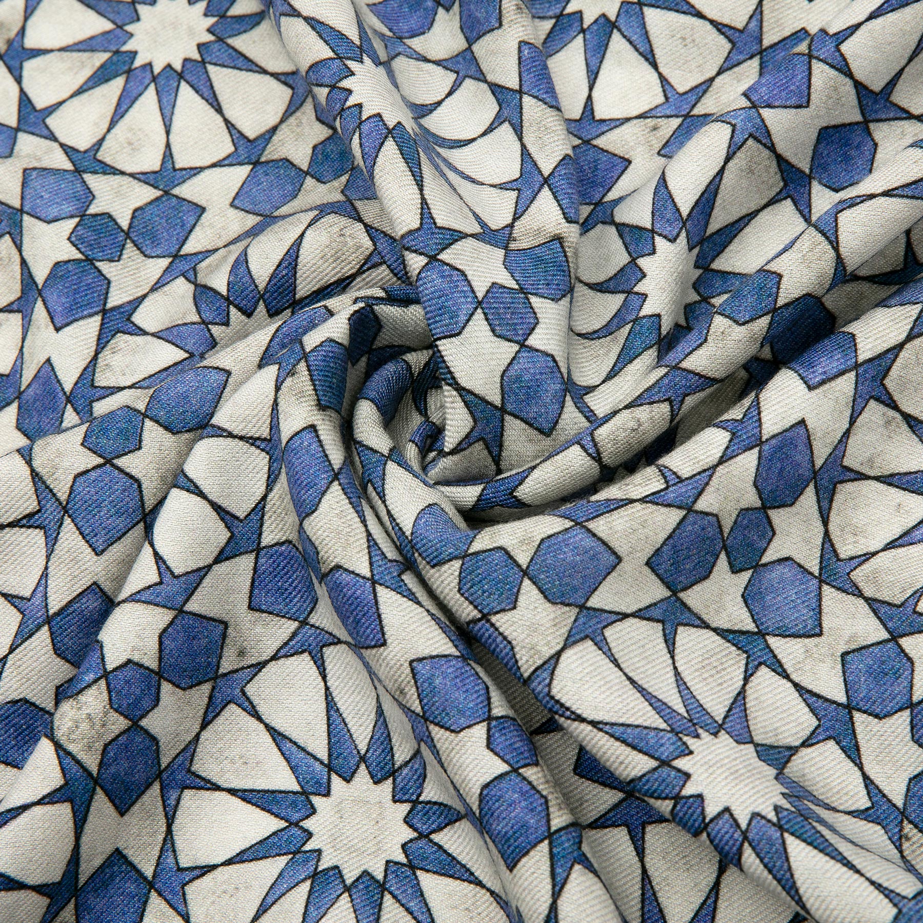 A close-up of a Silk and Wool Scarf with Blue and Gray Print shows interlocking geometric shapes and symmetrical designs, capturing cultural artistry. The fabric’s visible folds and creases enhance its textured beauty.