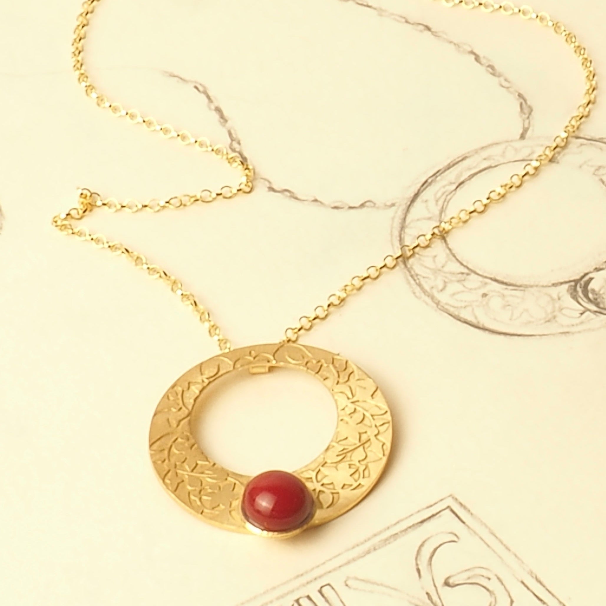 A Platónica Gold and Red Pendant inspired by Alhambra Palace, with floral engravings and a red stone, is shown on a cream background next to a partial pendant sketch.