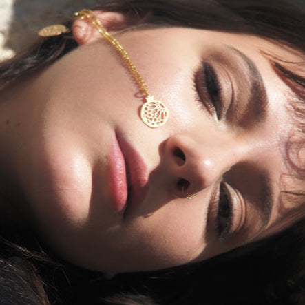 A woman with closed eyes lies down, wearing the Gold Plated Pendant Granada No.1 earring, its round, intricate design resting against her cheek as sunlight highlights her face and makeup.
