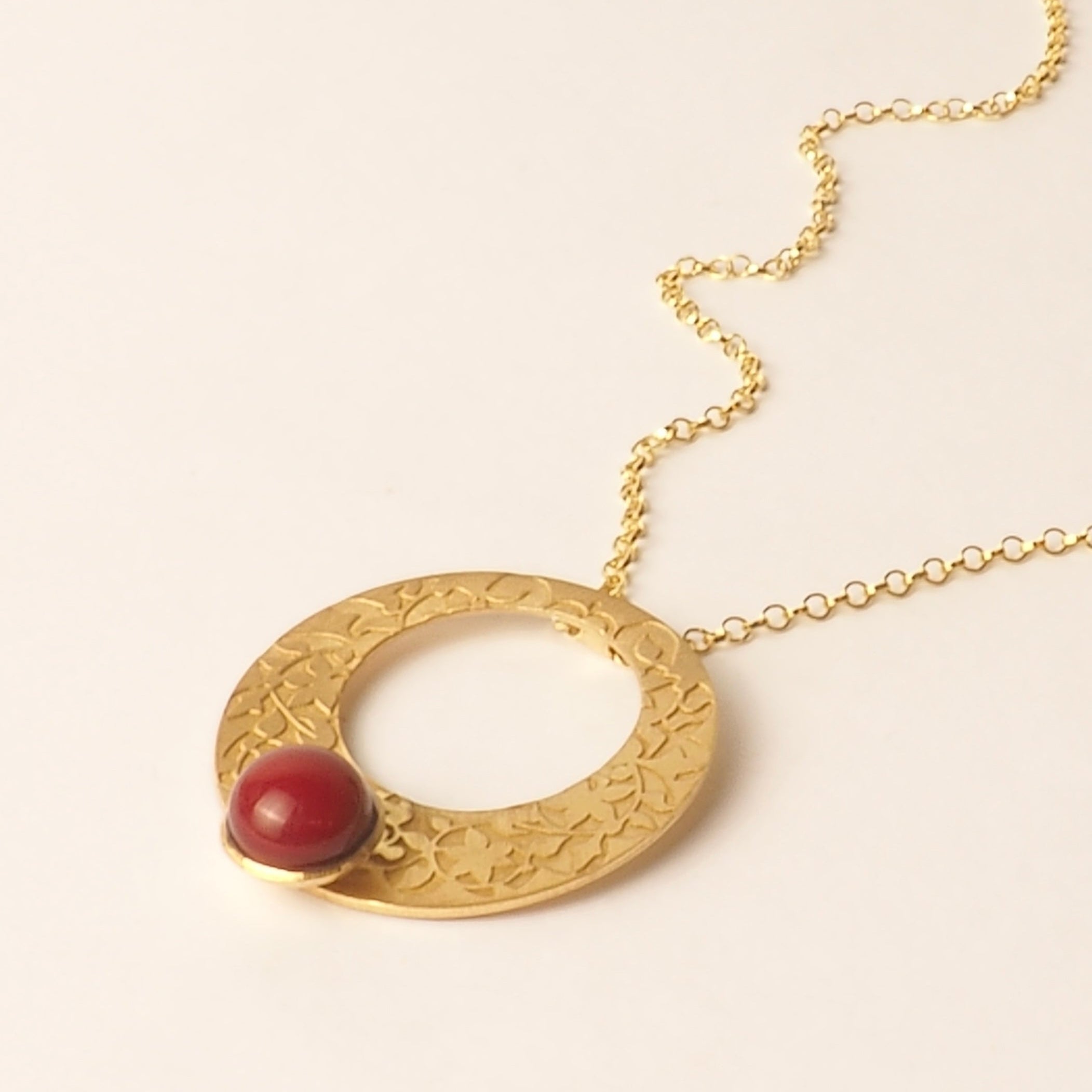 The Platónica Gold and Red Pendant, inspired by Alhambra Palace, features a textured circular charm with a vibrant red bead, shown on a light neutral background.