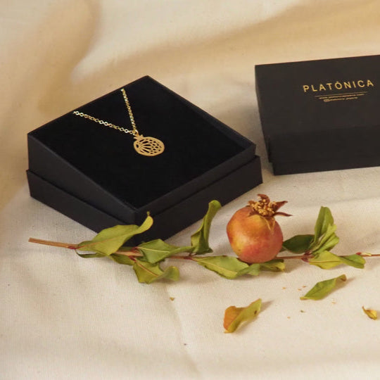 The Gold Plated Pendant Granada No.1, handcrafted in Spain, rests in a black jewelry box beside a branded Platónica box, accompanied by a pomegranate and leafy branch on cream-colored fabric.
