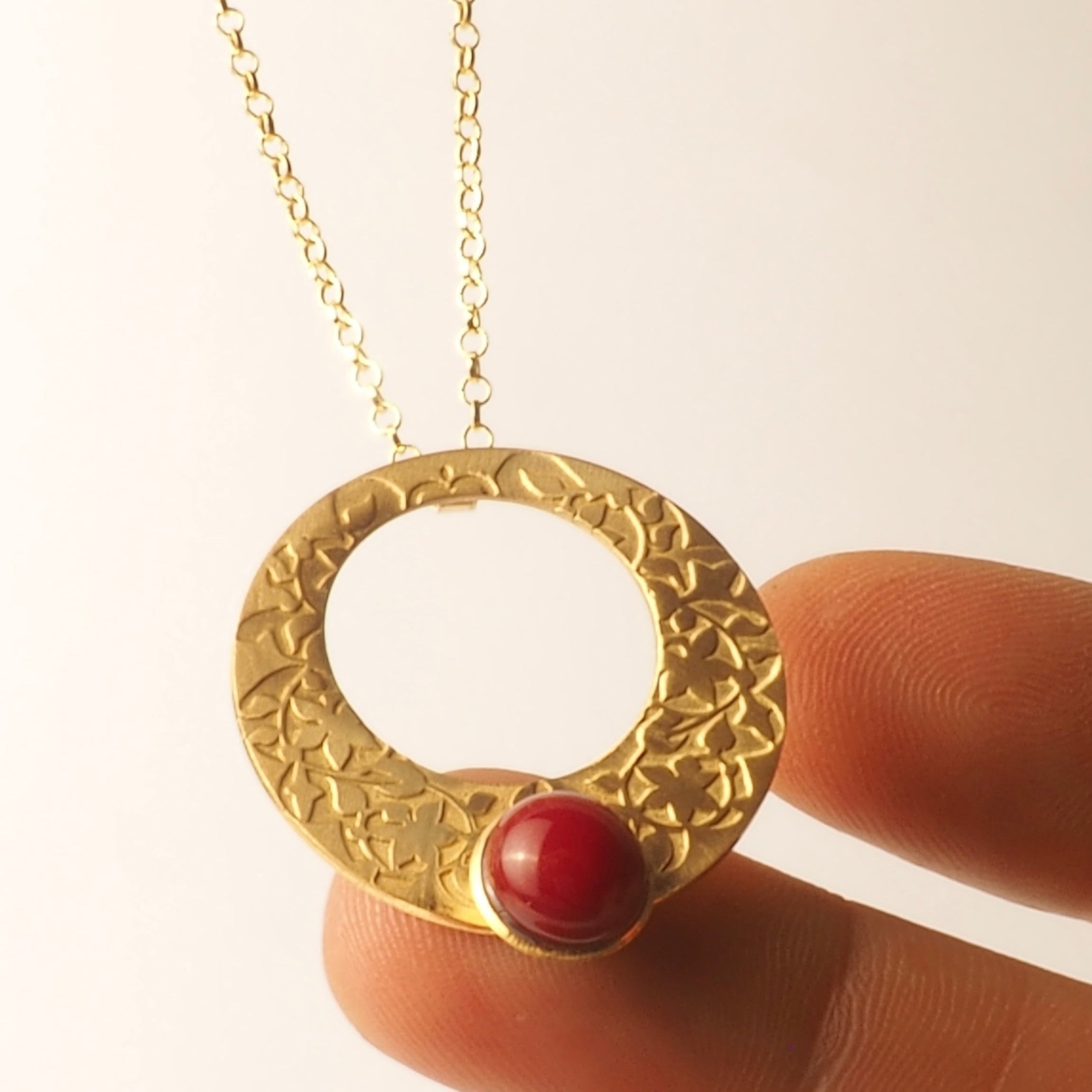 A hand holds the Platónica Gold and Red Pendant inspired by Alhambra Palace, with a circular, textured design and central red gemstone. The elegant necklace contrasts against a plain white background.