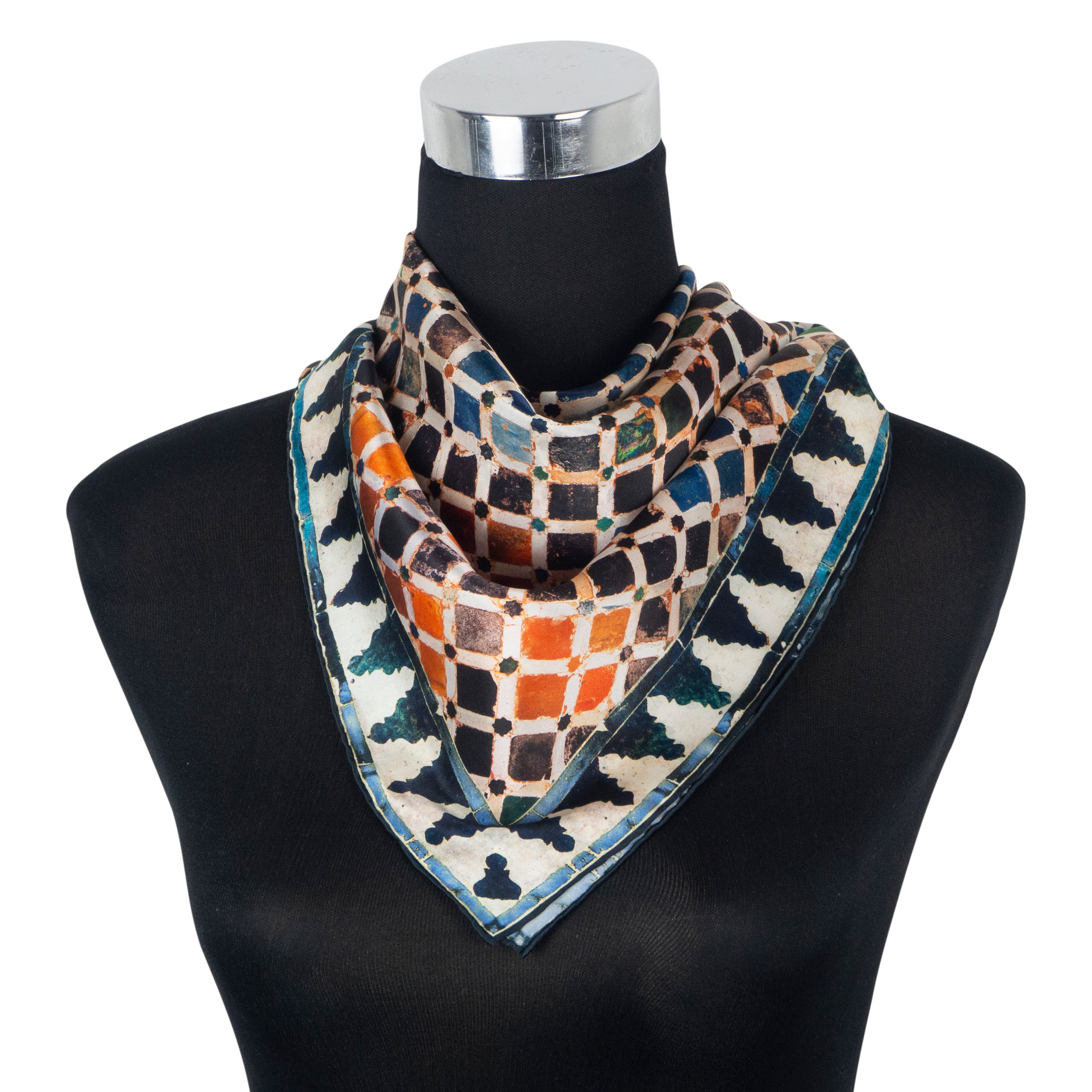 A black mannequin showcases the Multicolor Silk Square Scarf Comares, crafted from pure silk with a geometric print in black, cream, orange, and green. The scarf is folded in a triangle and elegantly draped around the neck, featuring hand-rolled hems.