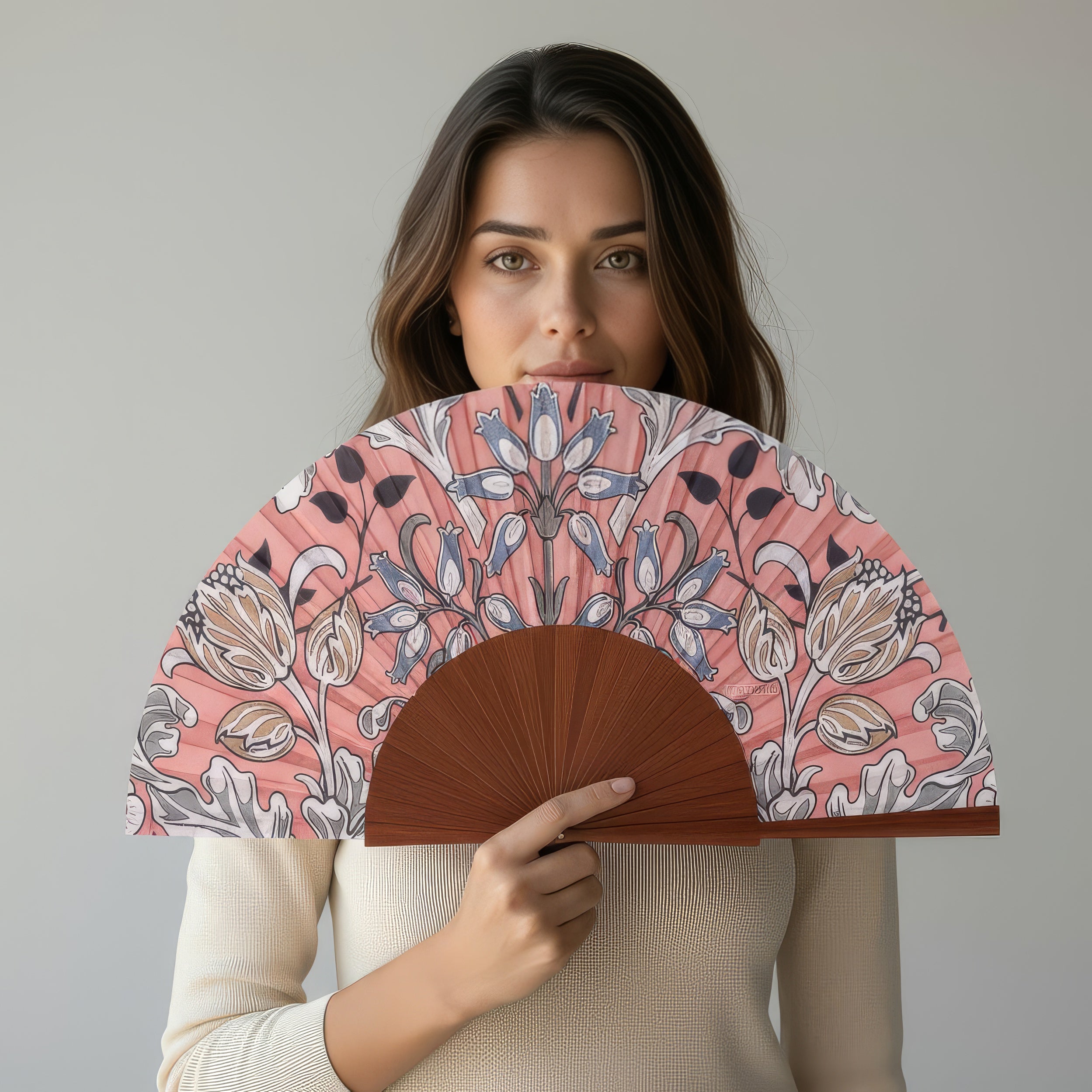 A woman with long brown hair, wearing a cream top, stands against a light gray background holding the Folding Silk Fan with Floral Print Elinor, partially covering her face.
