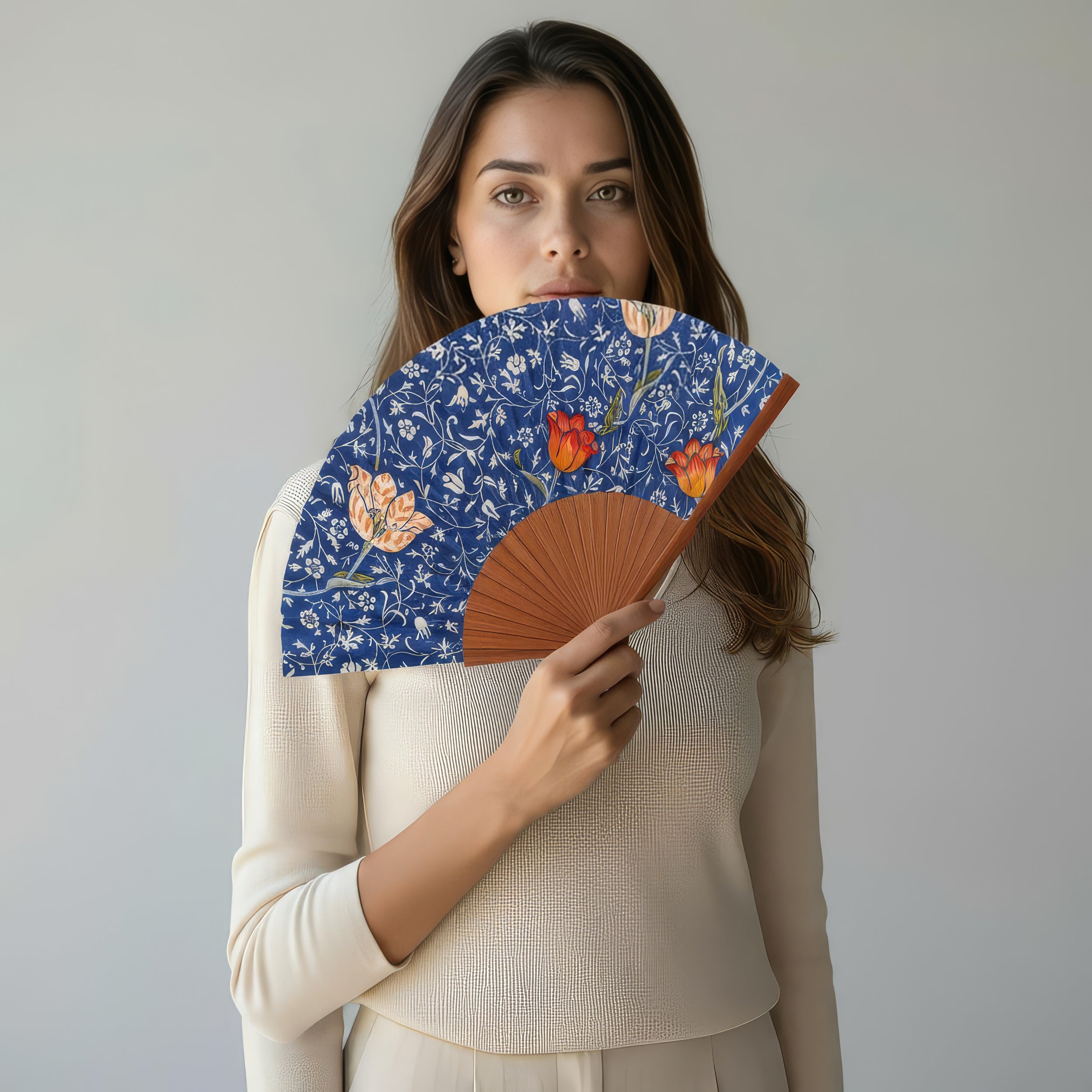 A woman with long brown hair in a cream top holds the Natural Silk Fan W. Morris Print Gálata, partially covering her face, highlighting Spanish craftsmanship against a plain light background.
