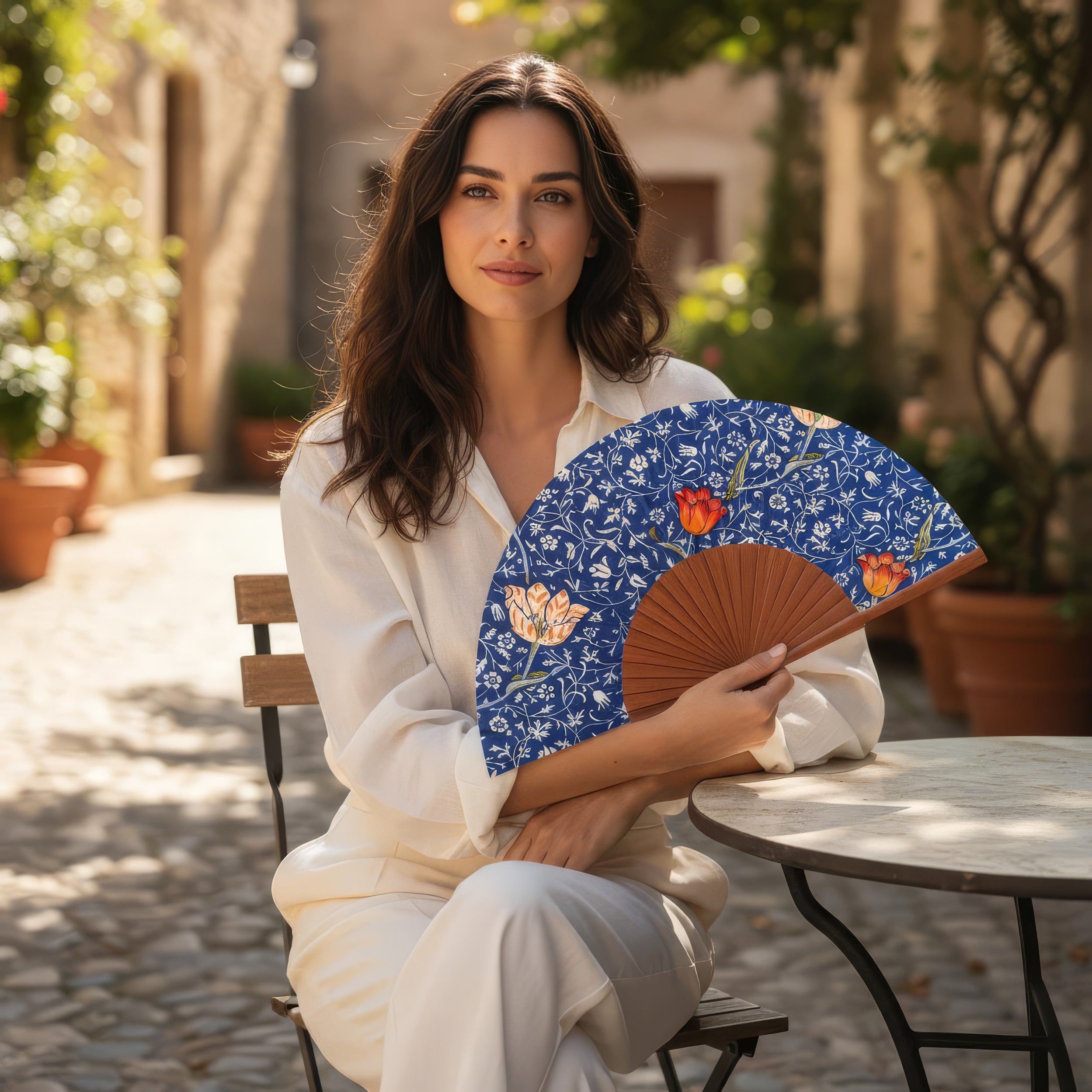 A woman in white sits at an outdoor café table, holding the Natural Silk Fan W. Morris Print Gálata. Sunlight filters through a cobblestone courtyard with potted plants in the background.