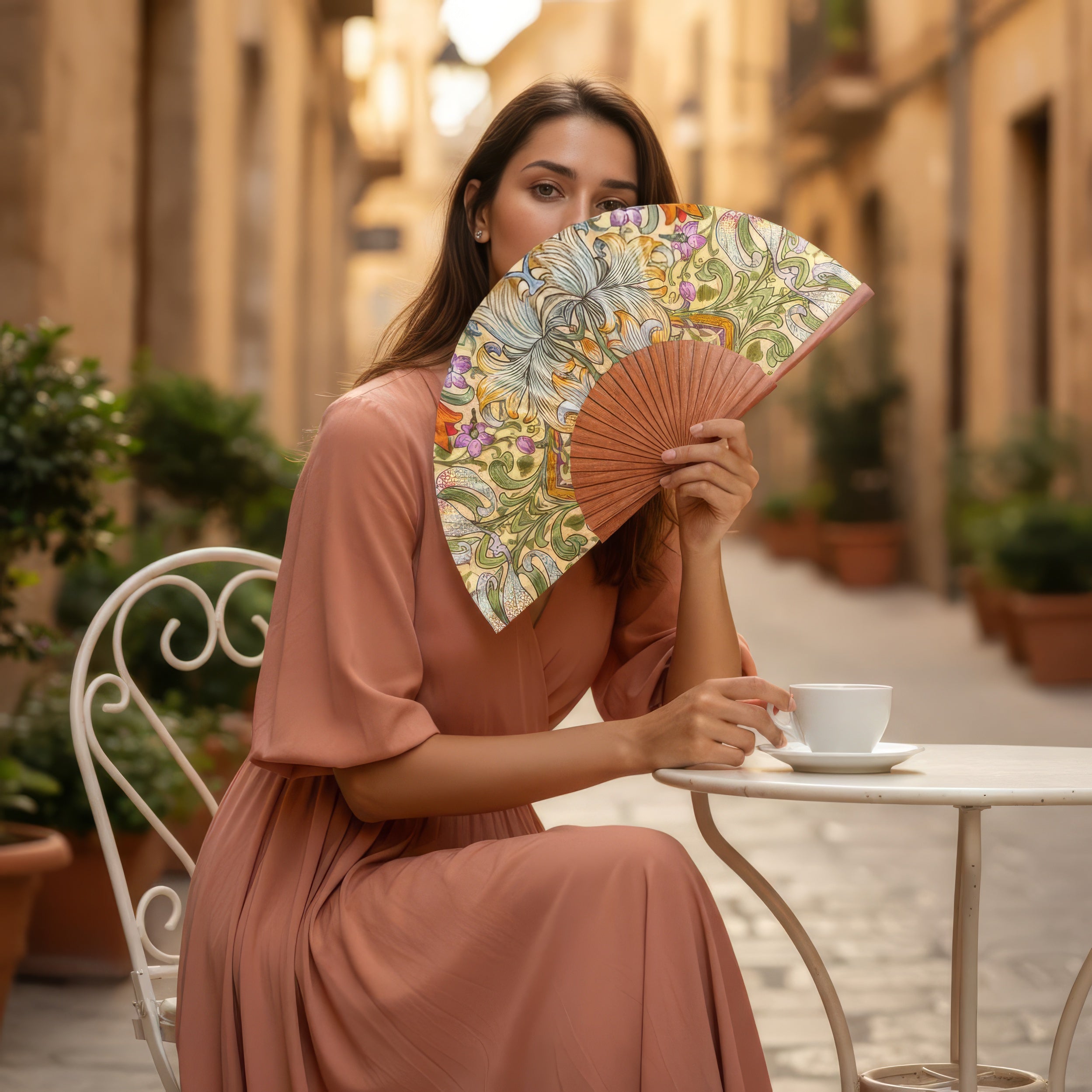 A woman in a flowing peach dress sits at an outdoor café, sipping coffee and holding the Spanish Hand Fan with Floral Print Georg, partially covering her face, on a sunlit street lined with potted plants.