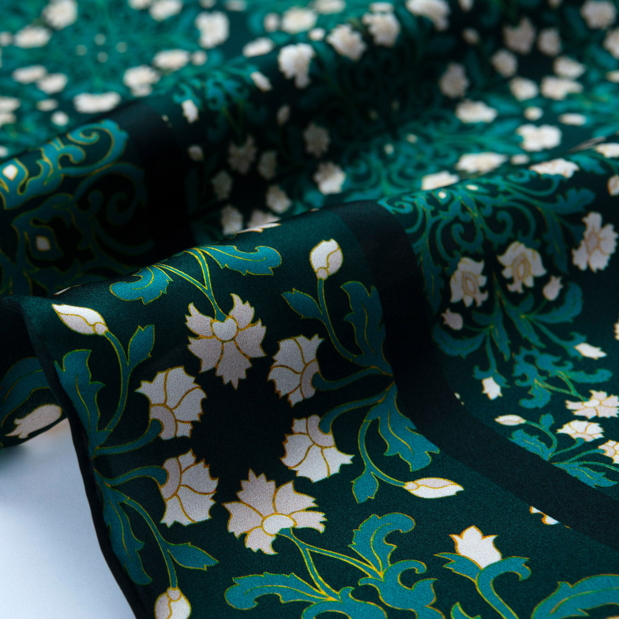 A close-up of the Floral Green Silk Scarf with Delicate White Blooms, draped to highlight its soft silk texture and intricate white floral pattern with swirling leaves in an art nouveau style.
