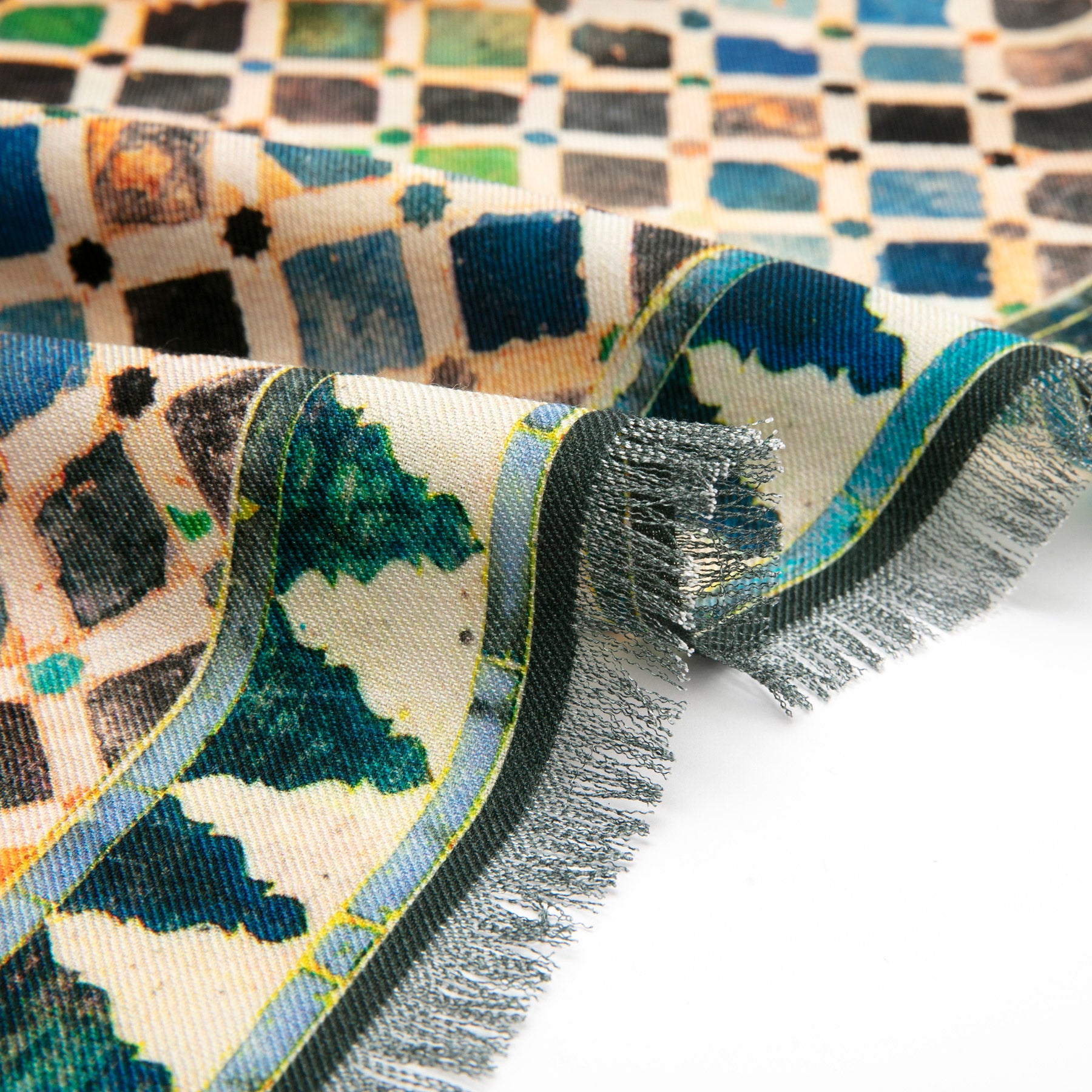A close-up of the Silk Wool Scarf Comares shows its vibrant mosaic geometric pattern with green, blue, black, and beige squares and frayed edges, set against a plain white background.