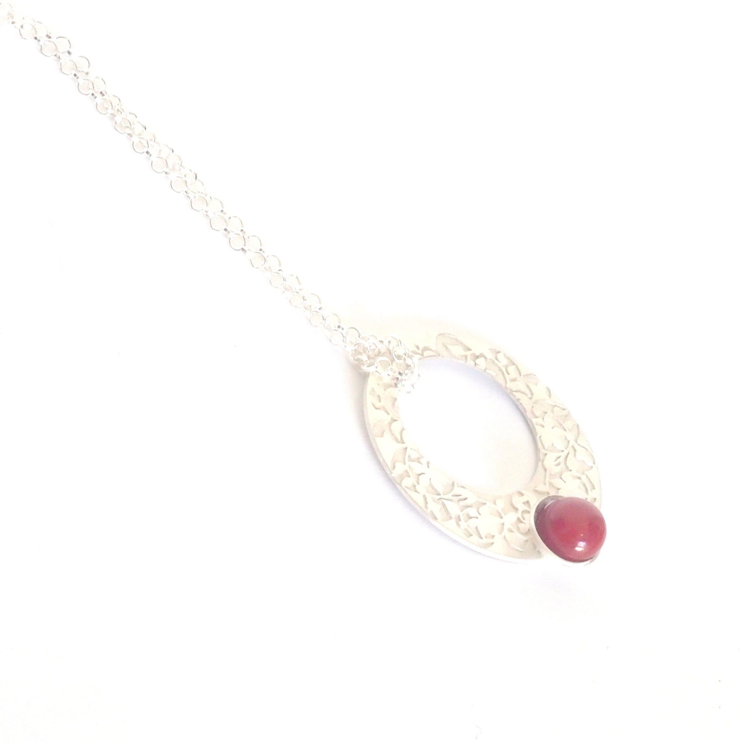 The Red Nasrid Palaces Detail Pendant by Platónica is a 925 silver necklace from the Alhambra collection. It showcases an oval pendant with a floral design, enhanced by a single red bead, and hangs elegantly on a slender chain against a pristine white background.