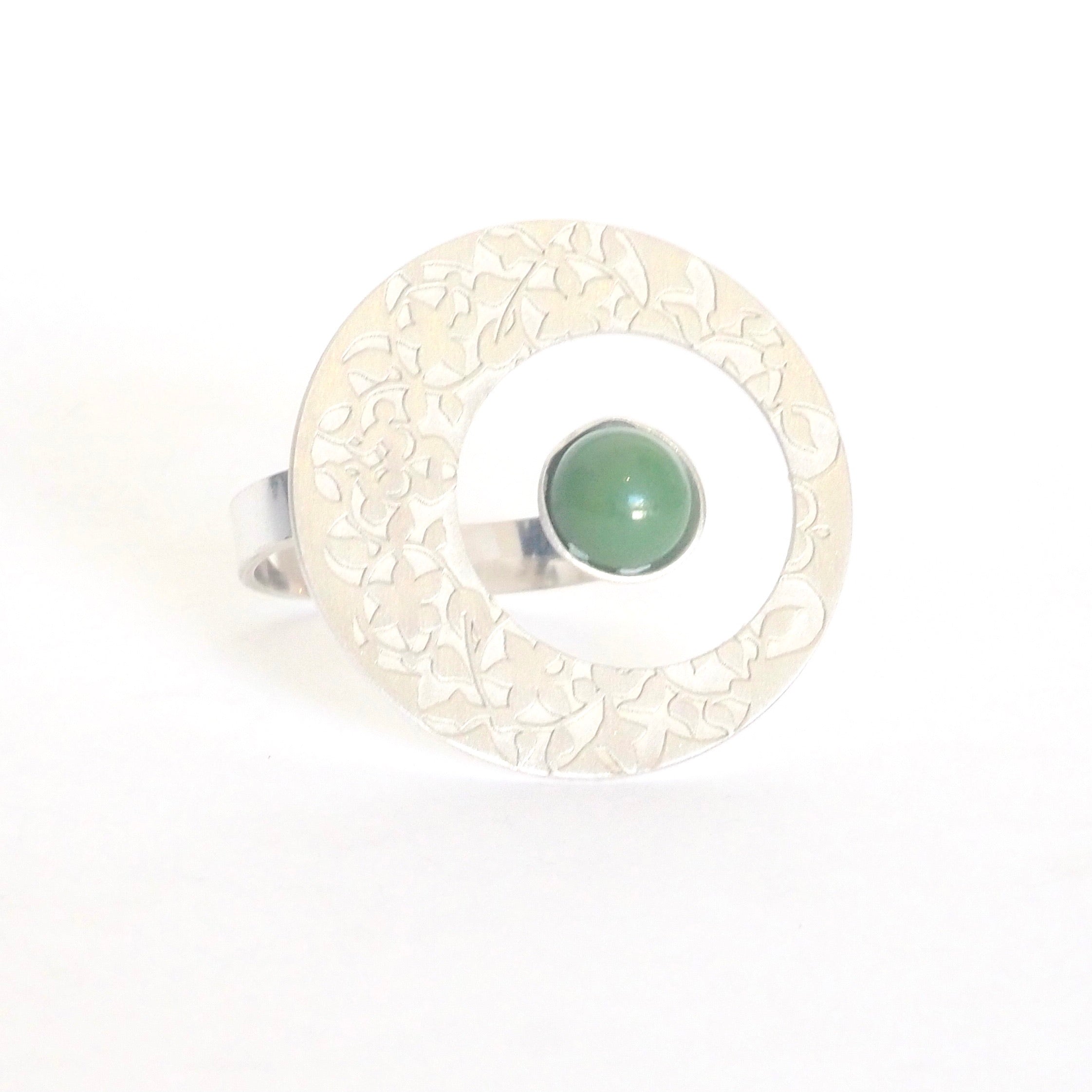 The Green Nasrid Palaces Circle Ring by Platónica, part of the Alhambra collection, is crafted from 925 silver. It has a round ornate disk with floral patterns and features a central green stone. The adjustable ring is elegantly presented upright against a plain white background.