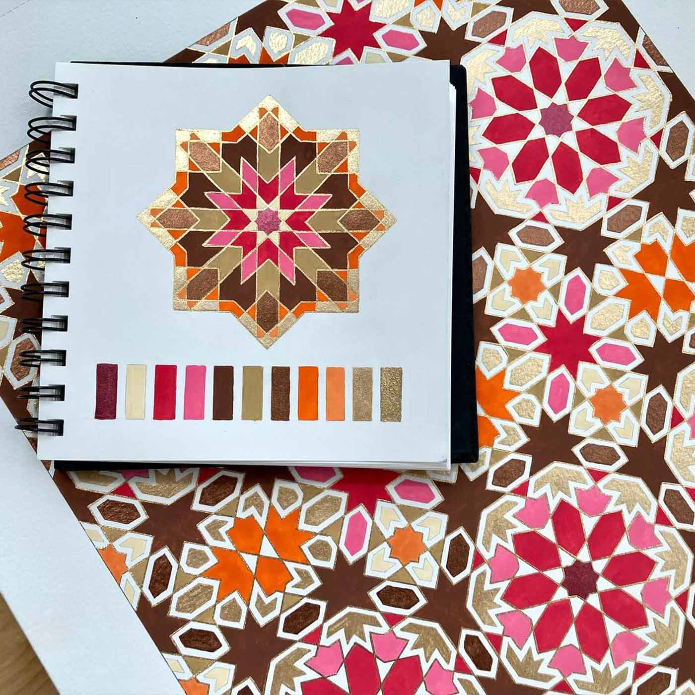 A sketchbook displays a geometric star pattern in brown, orange, red, pink, and gold, with matching color swatches below. The sketchbook rests on a sheet featuring a larger, similar repeating pattern.