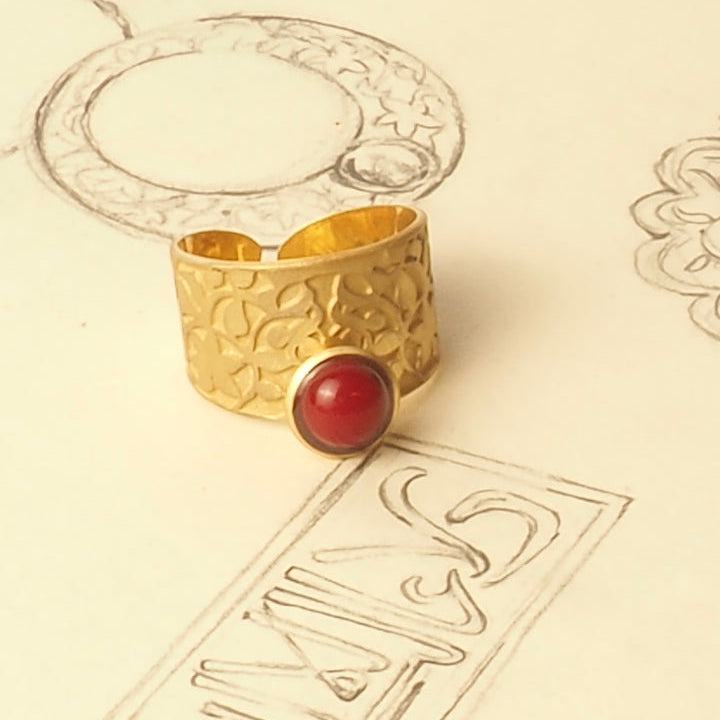 The Nasrid Palaces Detail Ring Red Gold by Platónica, featuring intricate patterns and a red gemstone, rests on paper with jewelry sketches that echo the exquisite design and artistry of the Alhambra collection.