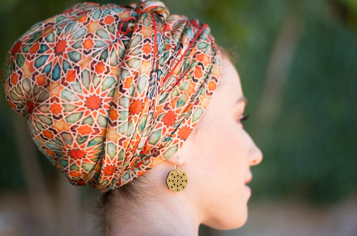 A woman in profile view wears a colorful patterned headscarf with geometric designs and a gold, intricate round earring. The background is blurred greenery.