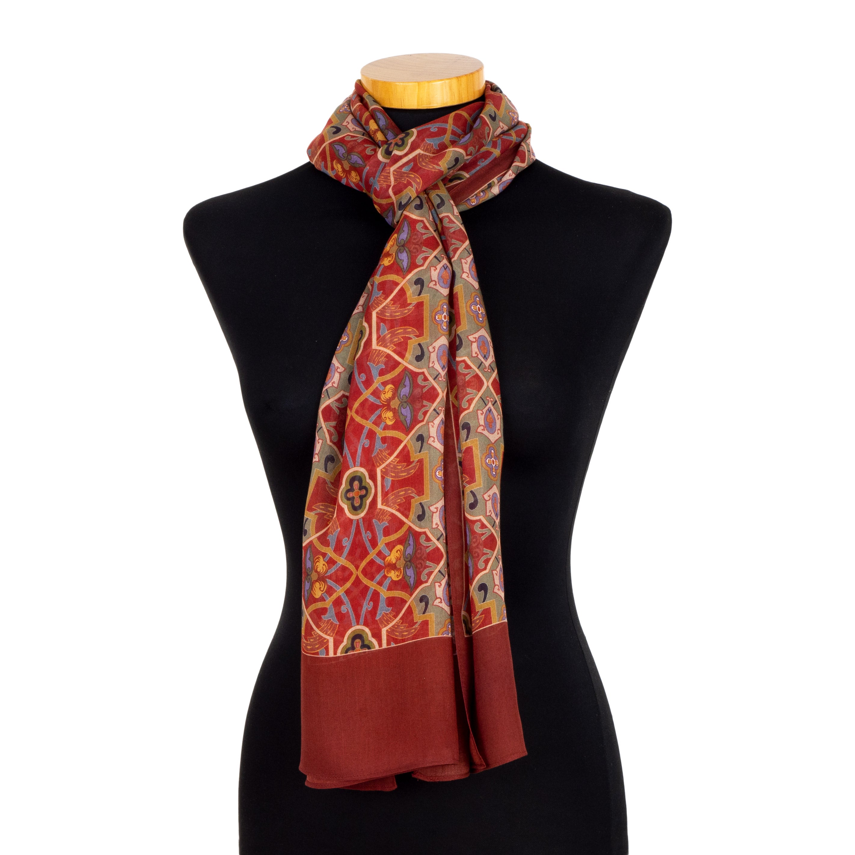 A mannequin displays the Red Scarf with Floral Print Samra, inspired by the Alhambra, featuring orange and gold geometric and floral motifs on red, with a solid dark red border and end, loosely wrapped around the neck.