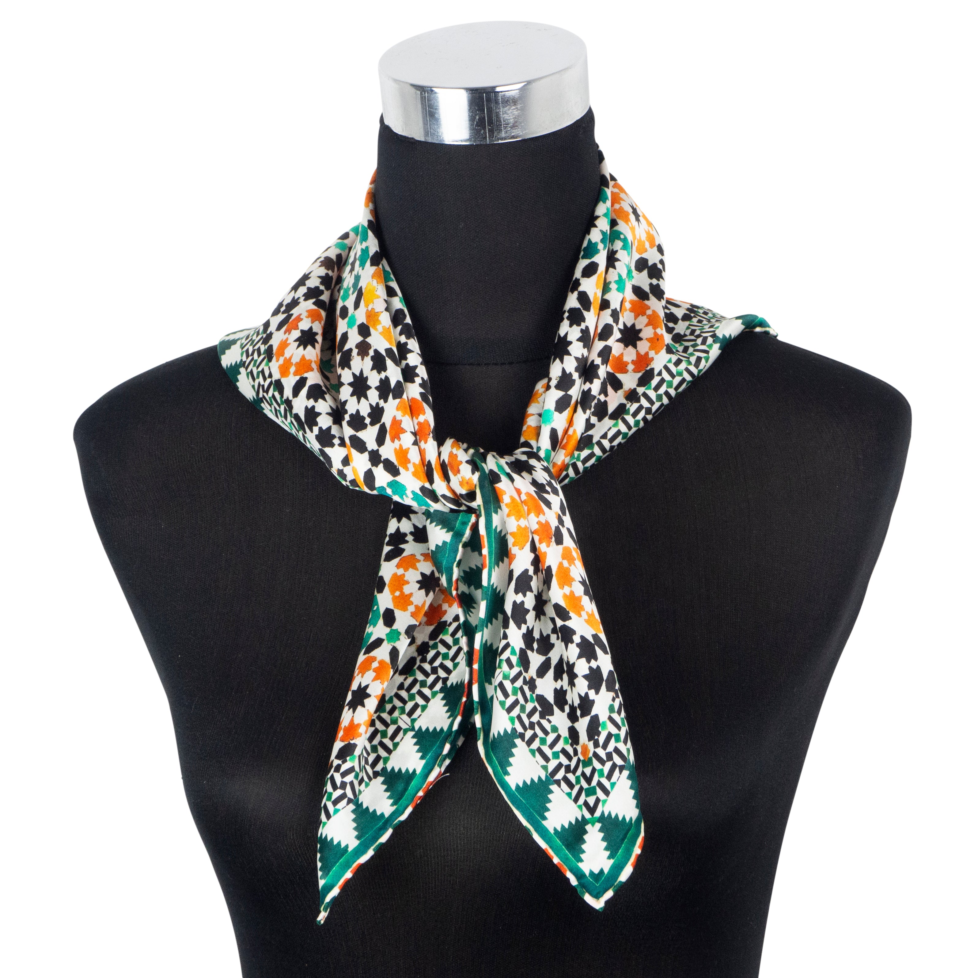 A mannequin showcases the Islamic Art Square Silk Scarf Sabika Green, featuring geometric and floral patterns in black, white, orange, and green, tied at the neck with hand rolled edges.