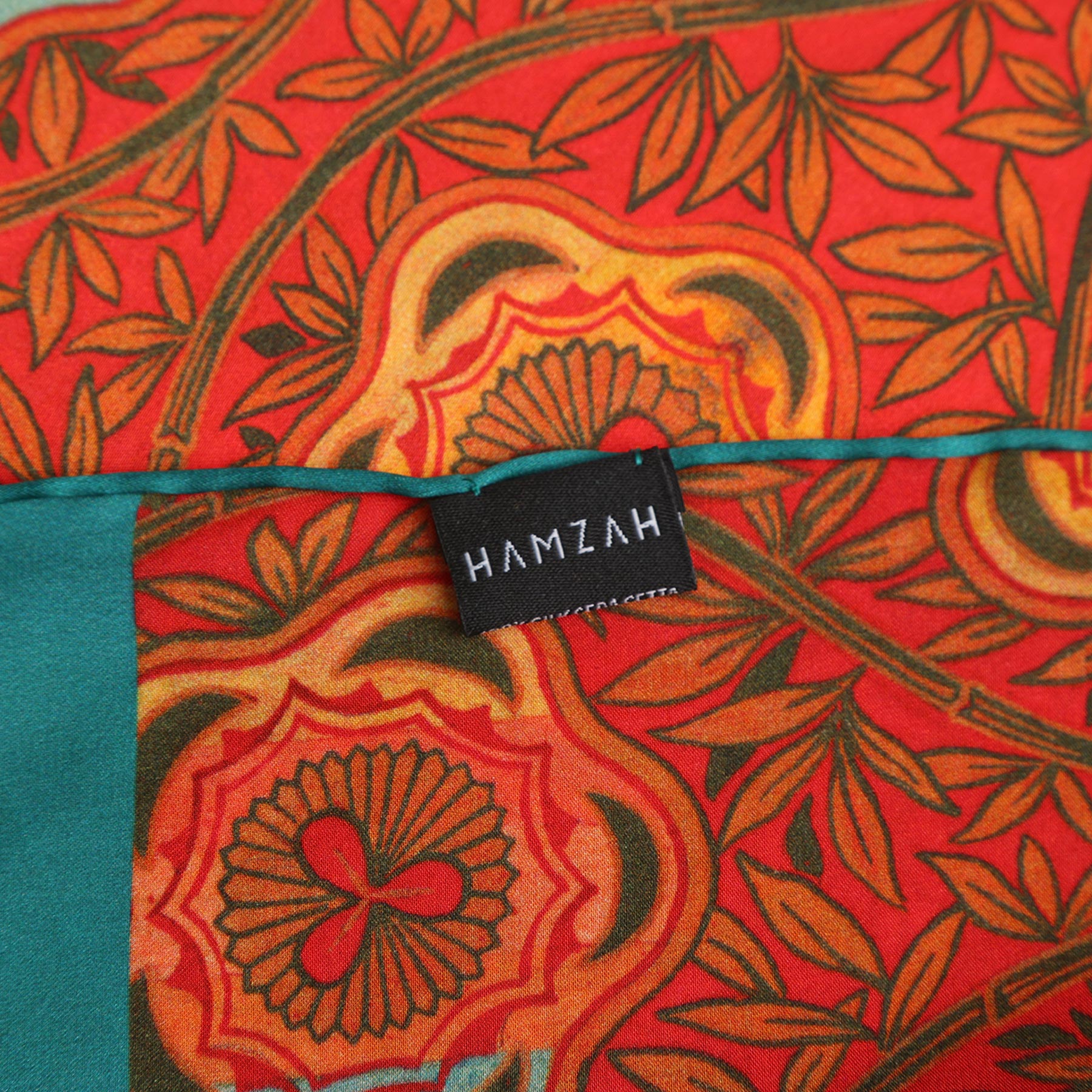A rectangular black box with HAMZAH at the top features a window showcasing the Red and Blue Scarf with Floral Print Marigold. Wrap yourself in elegance is written at the bottom.
