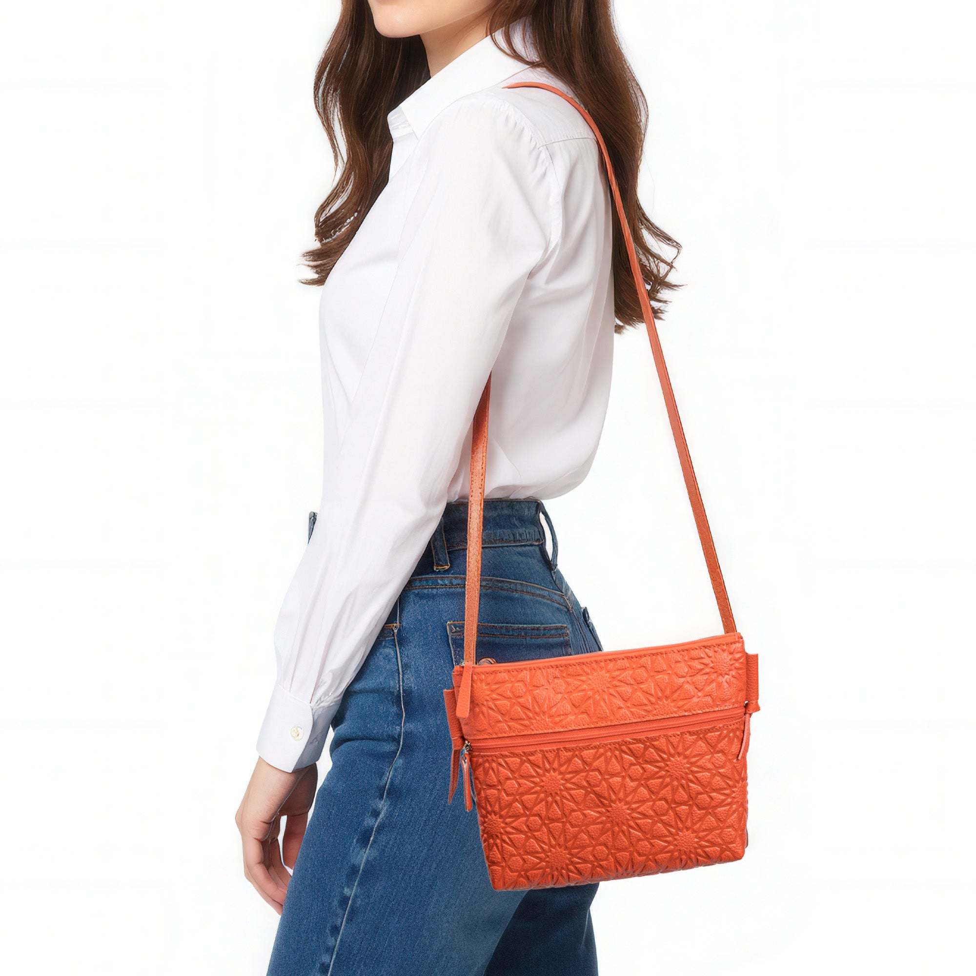 A woman in a white long-sleeve shirt and blue jeans carries an Orange Embossed Crossbody Bag with Islamic Design Pattern, standing against a plain white background.