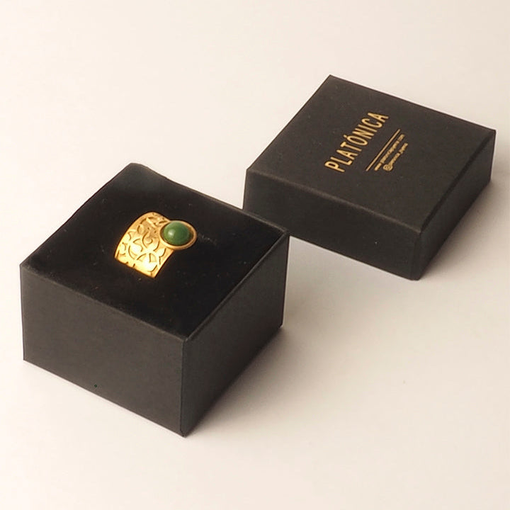 The Ring Inspired by Nasrid Motifs Gold & Green from Platónica, featuring an Alhambra-inspired engraving and a green stone, is displayed in an open black box beside a closed black PLATÓNICA box on a light surface.