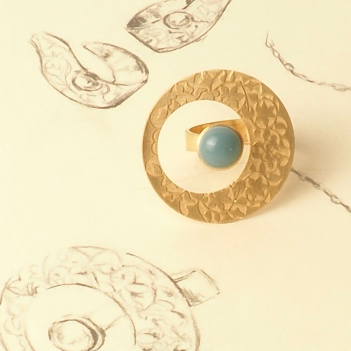 Platónicas Alhambra Palace-Inspired Gold Ring for Women, featuring a textured circular design with a blue detail, rests on paper showcasing sketched architectural designs reminiscent of the Alhambra Palace.