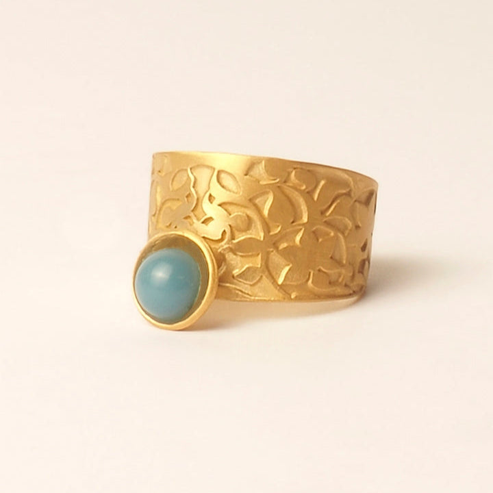 The Platónica Nasrid Palace Inspired Ring Gold & Blue features an embossed floral design with a smooth turquoise stone in a gold bezel on one side, showcased against a plain light background.
