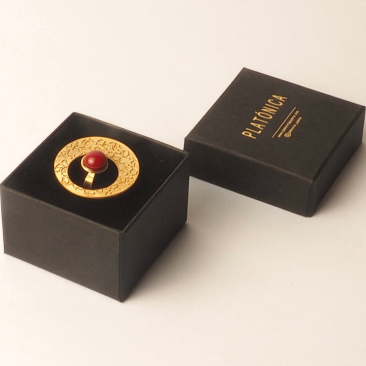 The Alhambra Palace inspired Gold Ring with Red Stone from Platónica is displayed on a black platform, alongside an open black box inscribed with PIKOMOA in gold, set against a light background.