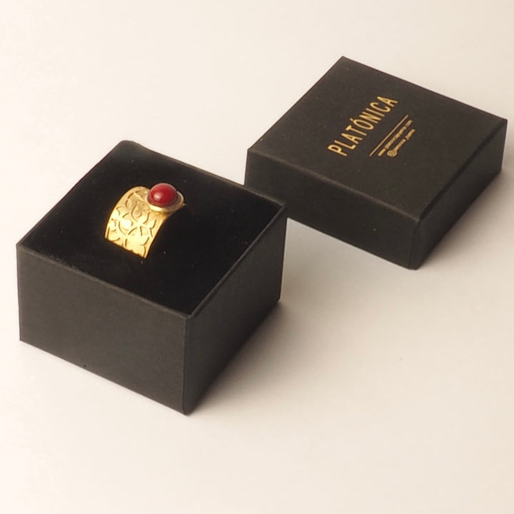 The Nasrid Palaces Detail Ring Red Gold by Platónica features a red gemstone in a gold setting, displayed in a black jewelry box with a closed Platónica box in gold lettering, reminiscent of the Alhambra collection, against a plain backdrop.