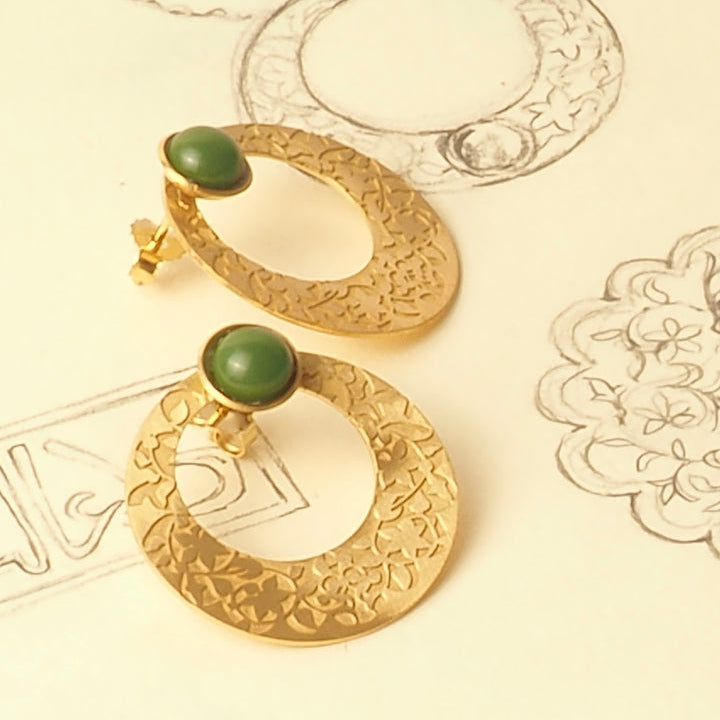 Platónicas Gold Earrings with Green Glass Stone, embossed with Alhambra Palace details, are elegantly displayed on paper featuring hand-drawn jewelry design sketches.