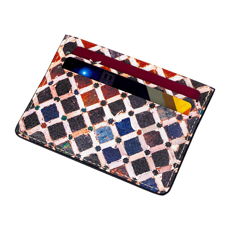 The Munira Leather Cardholder Comares is a multicolored, patterned accessory featuring a diamond grid design and holds three cards—a red, a gold, and a blue and white card—shown against a white background.