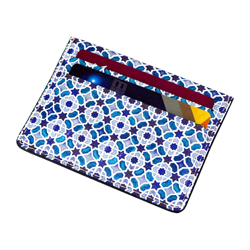 The Munira Leather Cardholder Alcázar is a blue and white leather accessory with a geometric pattern inspired by the Alcázar de Sevilla, featuring two slots for colorful cards, including a yellow-striped one.