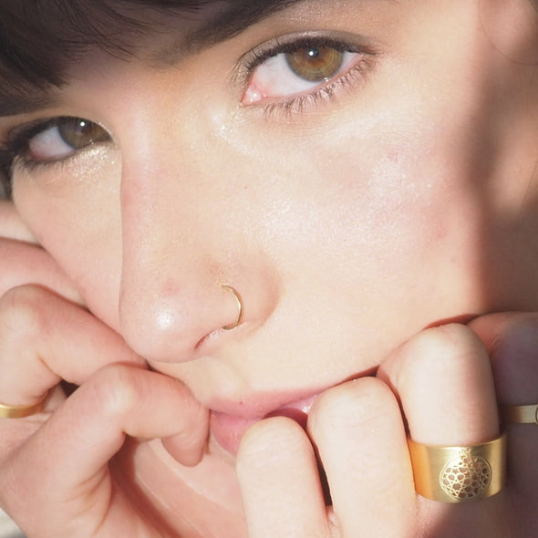 Close-up of a person with a thoughtful expression, wearing the Gold Plated Wide Ring Granada No.4 by Platónica on their finger. The lighting highlights their eyes and skin, creating a serene atmosphere.