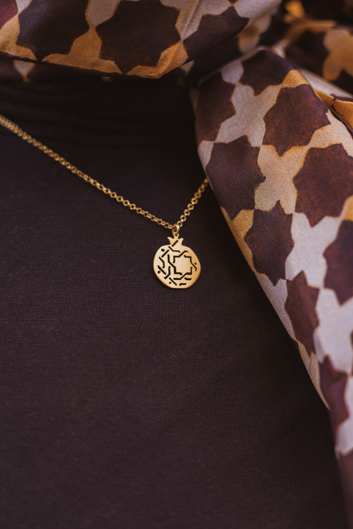 The Gold Plated Pendant Granada No.3 by Platónica, featuring an abstract design with lines and small shapes, hangs on a delicate chain against a dark fabric backdrop, beside a draped scarf in brown and beige geometric patterns.