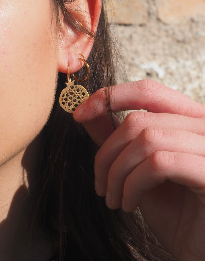 Someone gently holds Platónicas Gold Plated Hook Earrings Granada No.4, featuring a filigree design inspired by pomegranate branches, near their ear. A softly blurred brick wall background enhances the focus on the earrings intricate details and the persons hand.